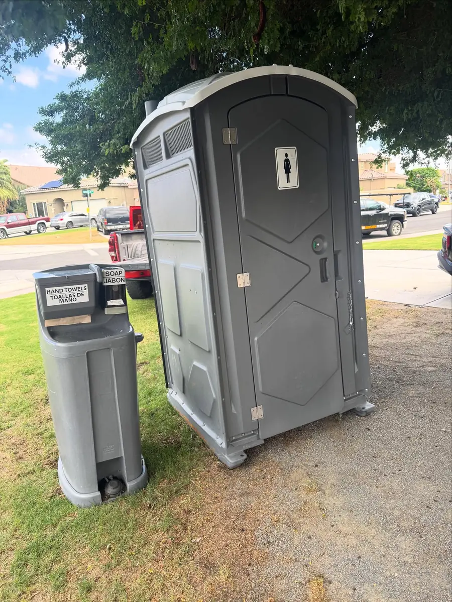 Gray standard porta potty with a hand-wash station set on a lawn beside a driveway